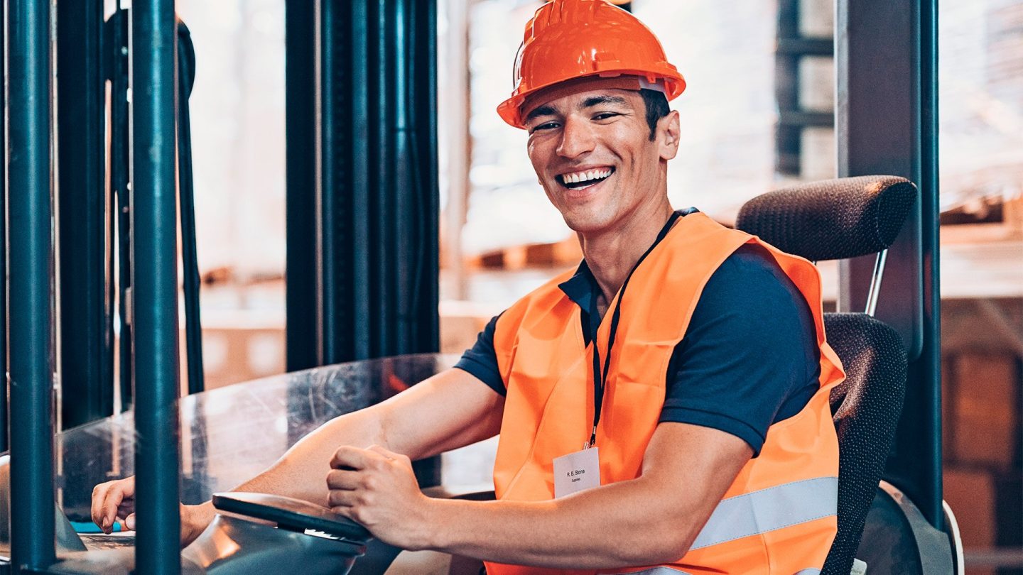 A man in an orange vest and safety helmet operating a forklift at a construction site.
