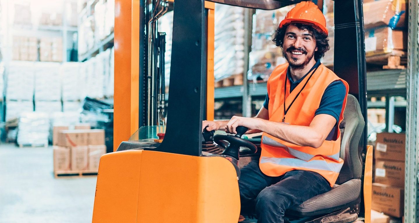 A forklift driver wears an orange hard hat and orange safety vest while driving an orange forklift