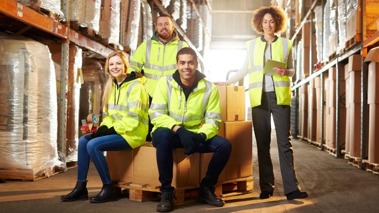 A group of young workers in a warehouse surrounded by cardboard boxes wearing safety jackets.