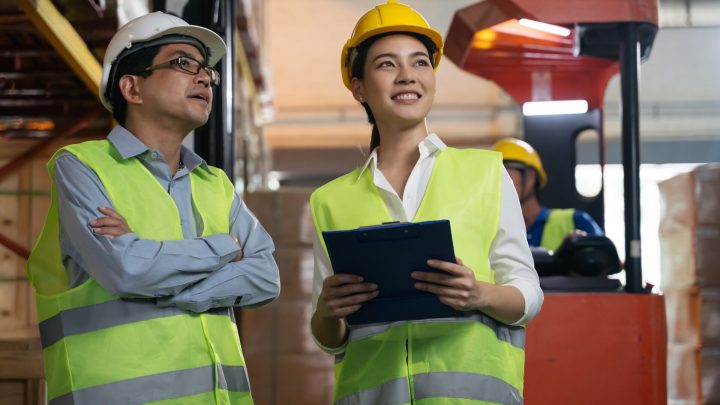 A female supervisor and male worker working in a warehouse surrounded by cardboard boxes wearing safety vests and hard hats.