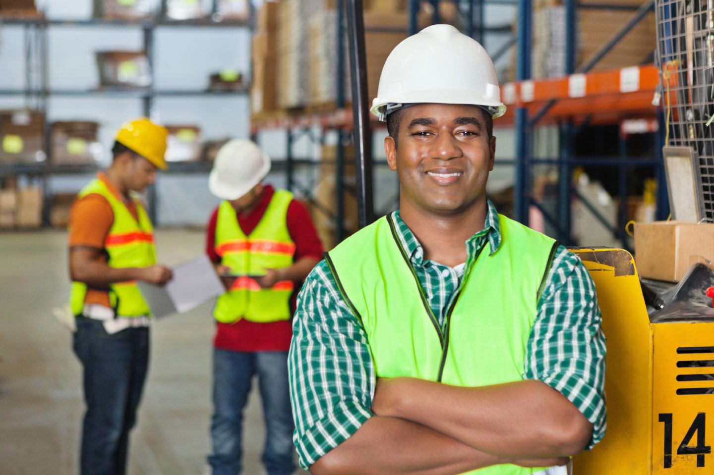 A young man is smiling at the camera with his arms crossed as he wears a hard hat and hi vis vest.