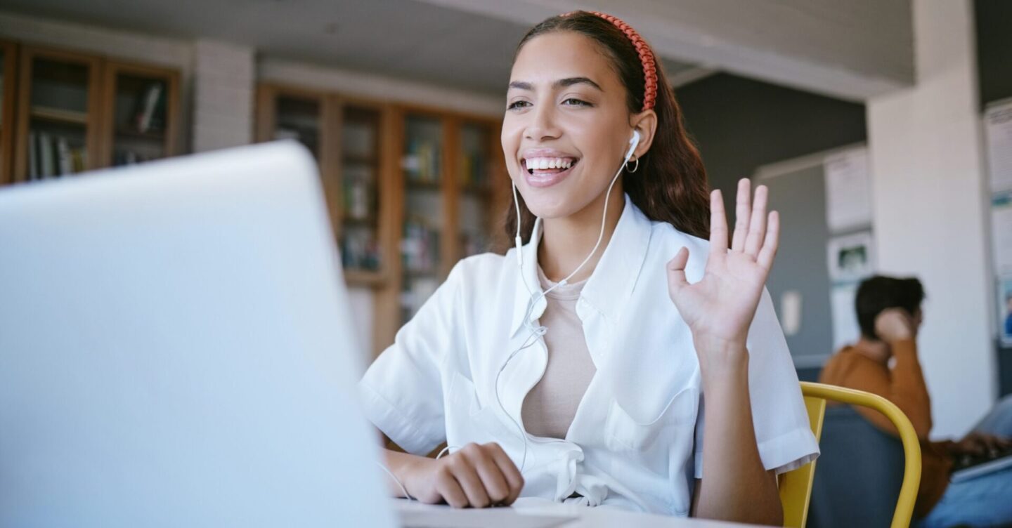 a smiling woman is sitting at a desk waving to her laptop screen for an online interview